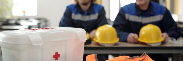 Close-up of table with first aid kit for factory workers, orange vest and respiratory mask, men with hardhats in background